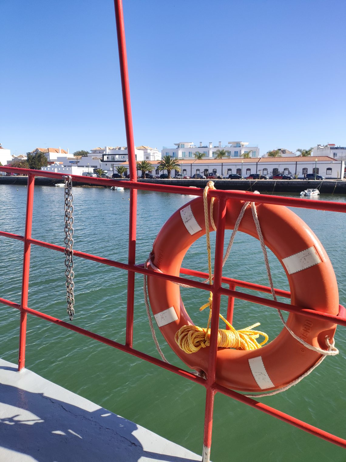 Orange lifebuoy secured to a red boat railing over calm harbor water with white buildings along the quay in the background.
