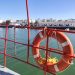 Orange lifebuoy secured to a red boat railing over calm harbor water with white buildings along the quay in the background.