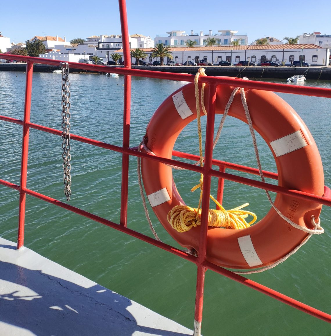 Orange lifebuoy secured to a red boat railing over calm harbor water with white buildings along the quay in the background.
