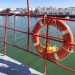 Orange lifebuoy secured to a red boat railing over calm harbor water with white buildings along the quay in the background.