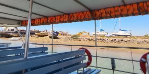 Deck seating on a boat with orange life jackets lining the canopy, overlooking a rocky shore with moored boats and clear blue water.